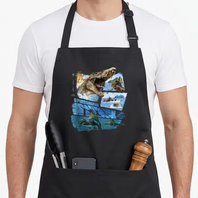 Attack spinosaurus apron - Spinosaurus head and boat collage with blue SPINOSAURUS RESEARCH text — shown on a black cotton canvas apron against a white background