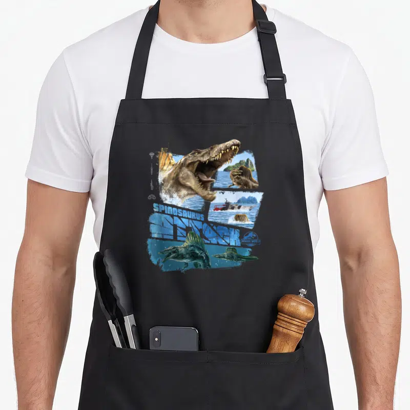 Attack spinosaurus apron - Spinosaurus head and boat collage with blue SPINOSAURUS RESEARCH text — shown on a black cotton canvas apron against a white background