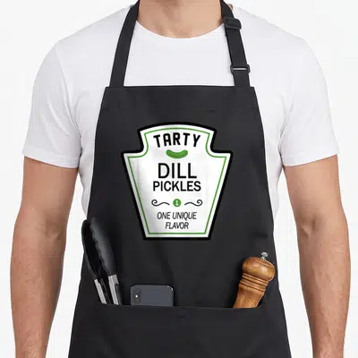 Dill halloween apron - TARTY DILL PICKLES text with green pickle icon and ONE UNIQUE FLAVOR in shield border — shown on a black cotton canvas apron against a white background