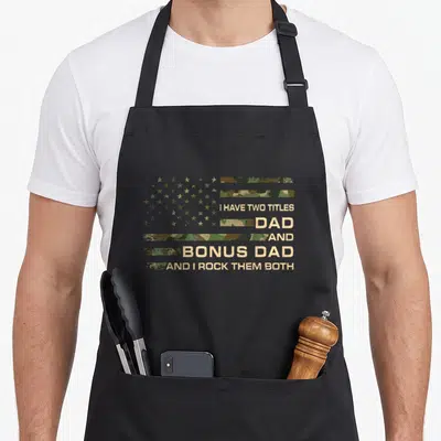 Title bonus father day apron - Distressed camo flag with text I Have Two Titles Dad Bonus Dad And I Rock Them Both — shown on a black cotton canvas apron against a white background