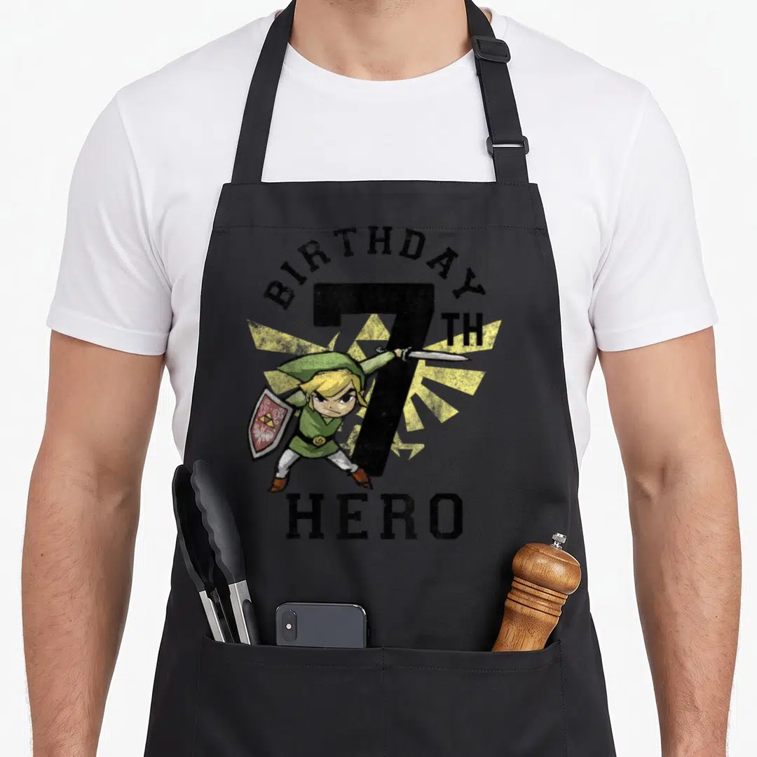Link birthday apron - Link holding sword and shield in front of number 7 with BIRTHDAY HERO text and yellow sunburst — shown on a black cotton canvas apron against a white background