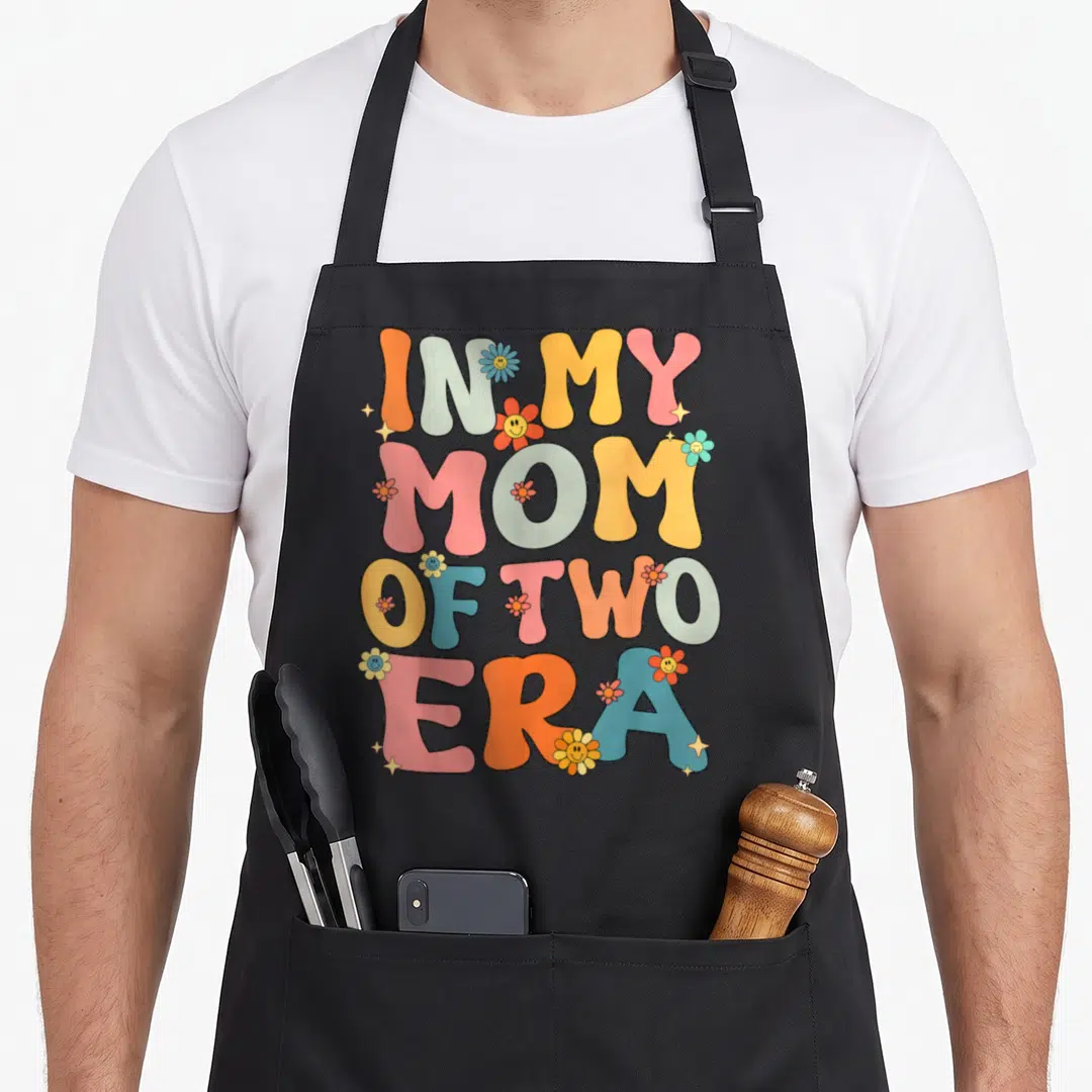 Era groovy mother's day aprons - Retro colorful text In My Mom Of Two Era with smiley face flowers and sparkles — shown on a black cotton canvas apron against a white background