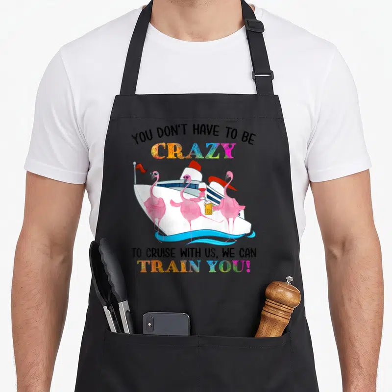 Cruise summer flamingo apron - Three pink flamingos holding wine and beer in front of a white cruise ship with colorful — shown on a black cotton canvas apron against a white background