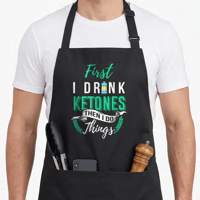 Drink lifestyle apron - Stacked text First I Drink Ketones Then I Do with water bottle icon and laurel wreath — shown on a black cotton canvas apron against a white background