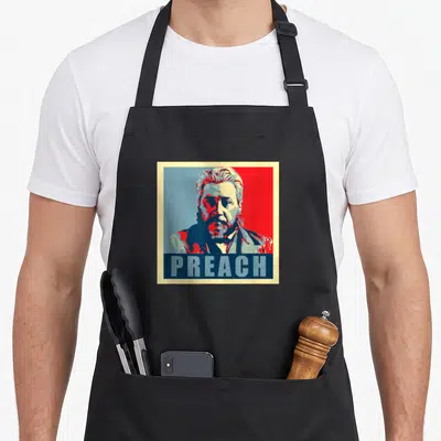 Product pilgrim apron - Stylized portrait of a bearded man above the word PREACH in red and blue — shown on a black cotton canvas apron against a white background