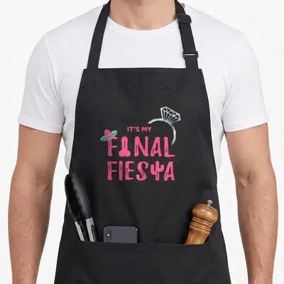 Final bride apron - Pink IT'S MY FINAL FIESTA text with diamond ring and sombrero graphic — shown on a black cotton canvas apron against a white background