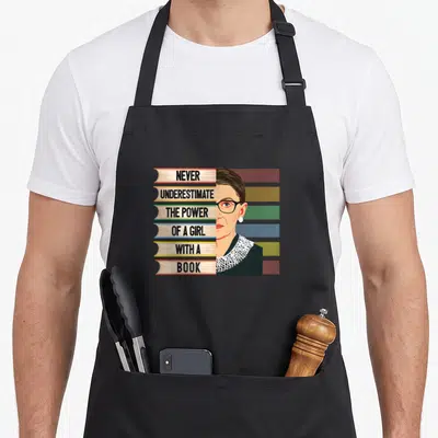 Feminist book quotes apron - Ruth Bader Ginsburg portrait next to six stacked books with bold text on spines — shown on a black cotton canvas apron against a white background