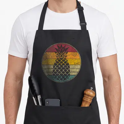 Fruit pineapple apron - Grey pineapple silhouette over red orange yellow teal horizontal stripes in distressed circle — shown on a black cotton canvas apron against a white background