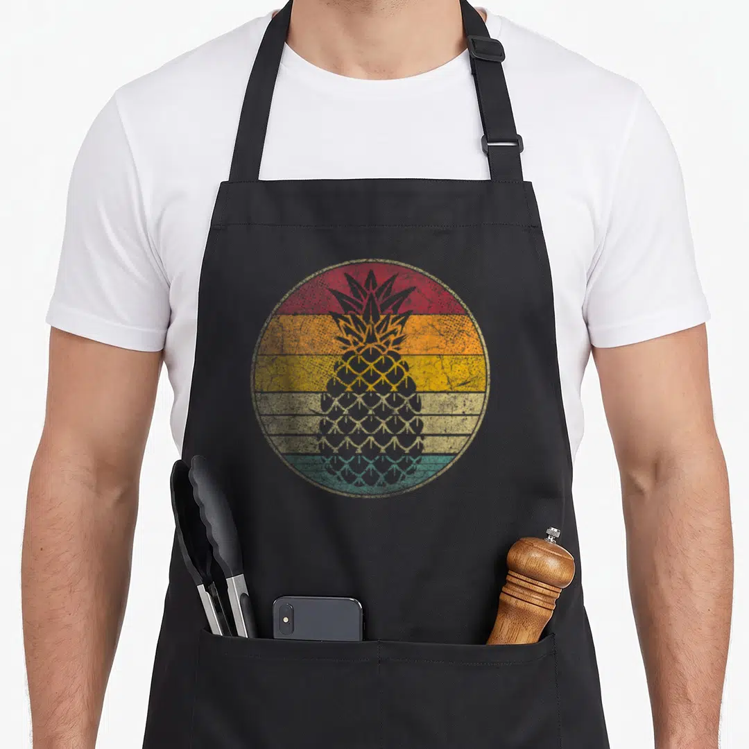 Fruit pineapple apron - Grey pineapple silhouette over red orange yellow teal horizontal stripes in distressed circle — shown on a black cotton canvas apron against a white background
