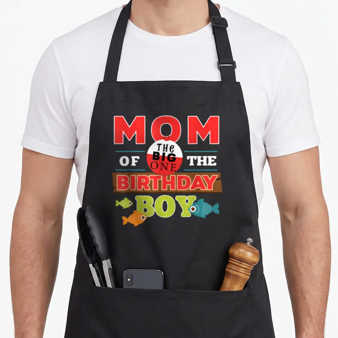 Theme fishing aprons - MOM THE BIG ONE OF THE BIRTHDAY BOY with bobber and three fish — shown on a black cotton canvas apron against a white background
