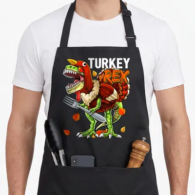 Rex turkey apron - Cartoon T-Rex in turkey costume holding fork with TURKEY REX text and scattered leaves — shown on a black cotton canvas apron against a white background