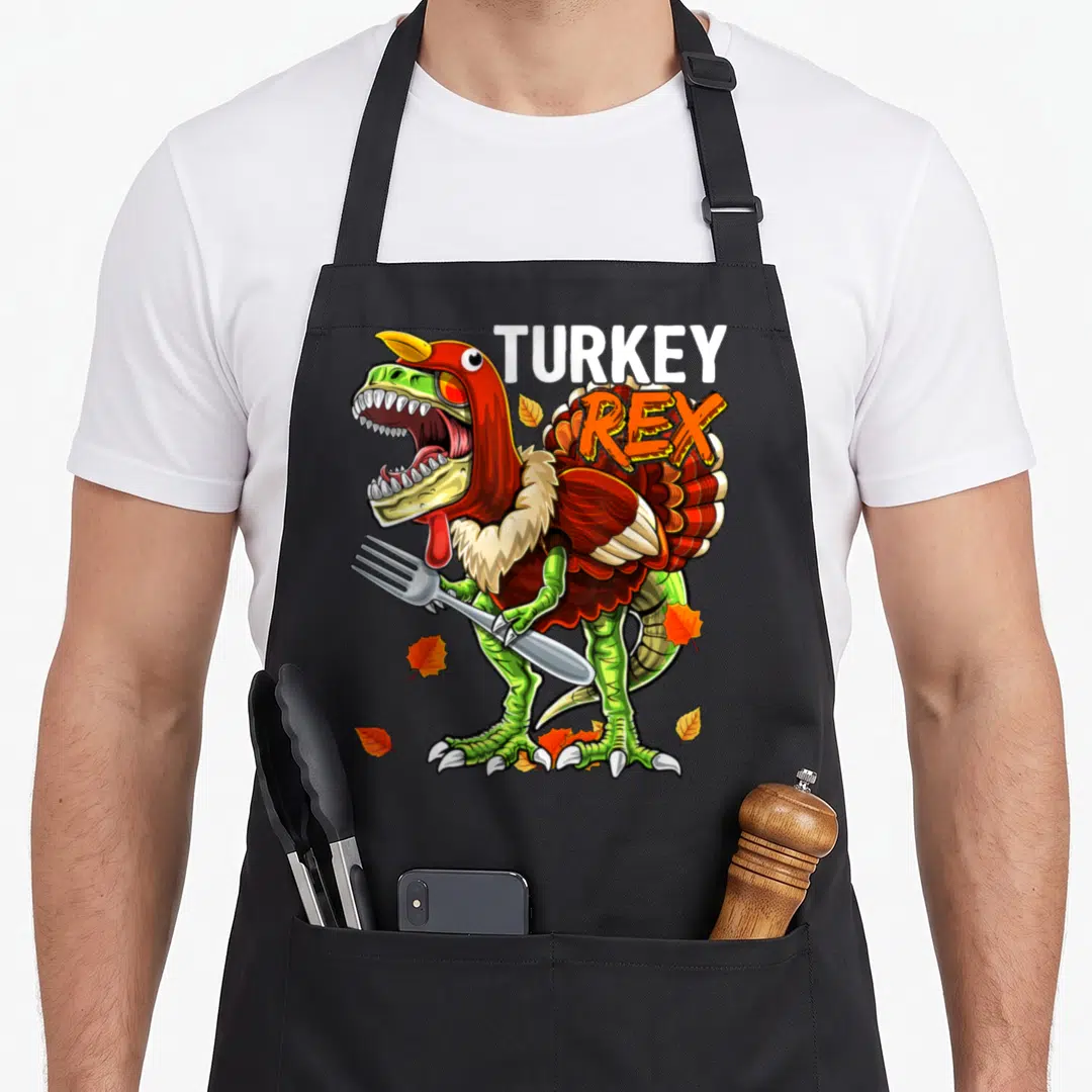 Rex turkey apron - Cartoon T-Rex in turkey costume holding fork with TURKEY REX text and scattered leaves — shown on a black cotton canvas apron against a white background