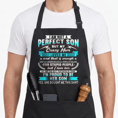 But mom and son aprons - I Am Not A Perfect Son But My Crazy Mom Loves Me text with ribbons and laurel wreath in white — shown on a black cotton canvas apron against a white background