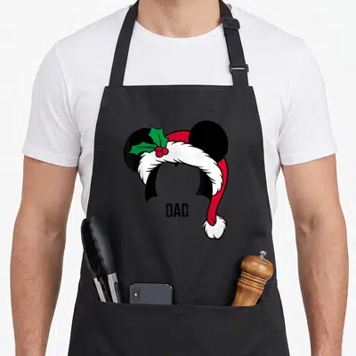 Ear holiday apron - DAD text below Mickey ears wearing Santa hat with green holly and red berries — shown on a black cotton canvas apron against a white background