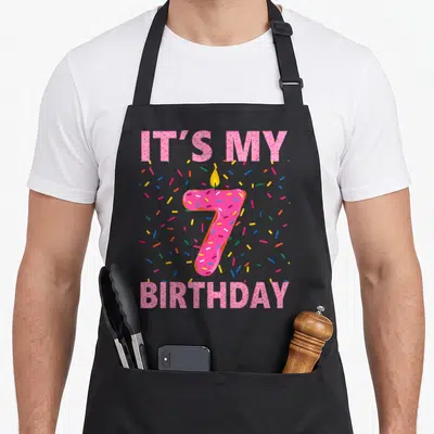 7th donut apron - Pink glazed number 7 candle with IT'S MY BIRTHDAY text and colorful sprinkles — shown on a black cotton canvas apron against a white background