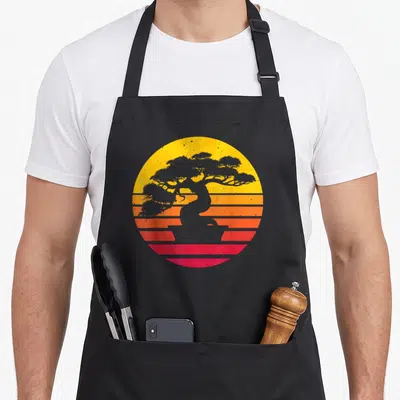Japanese culture tree apron - Grey bonsai silhouette on yellow orange and red striped retro circle — shown on a black cotton canvas apron against a white background