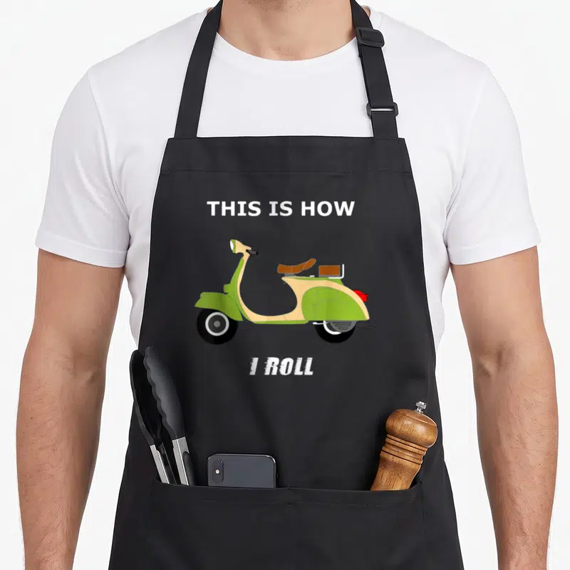 Moped italian apron - Green scooter illustration with This Is How I Roll text above and below — shown on a black cotton canvas apron against a white background
