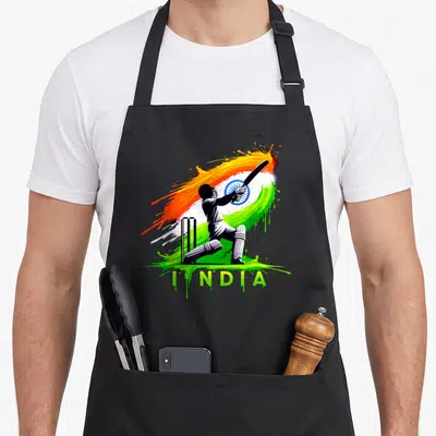 Fan cricket apron - Cricketer silhouette swinging bat over orange white green paint splatter with INDIA text — shown on a black cotton canvas apron against a white background