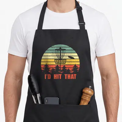 Hit golf apron - Retro disc golf basket with I'D HIT THAT text over striped sun and pine trees — shown on a black cotton canvas apron against a white background