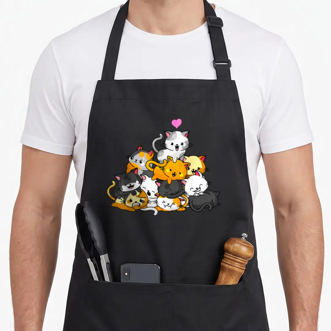Kitty kawaii cooking apron - Cartoon pile of orange, grey, white, black, and pink cats with a floating pink heart — shown on a black cotton canvas apron against a white background