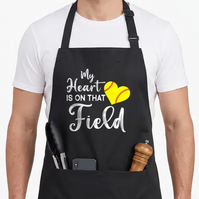 Field softball mom apron - My Heart IS ON THAT Field text next to yellow softball heart with red stitching — shown on a black cotton canvas apron against a white background