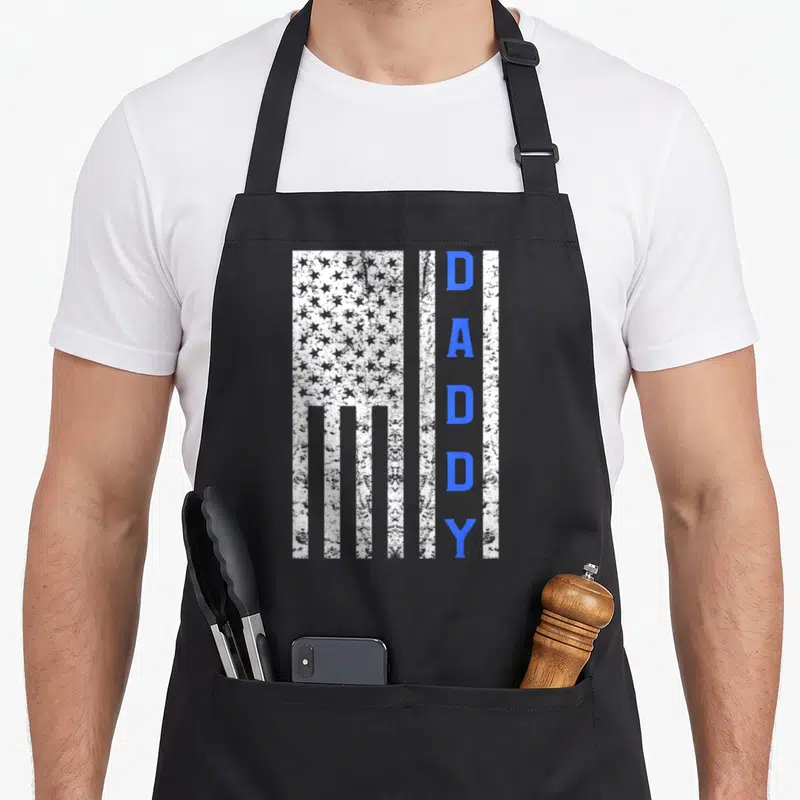 Cop father day apron - Distressed white vertical USA flag with blue vertical DADDY text on black apron — shown on a black cotton canvas apron against a white background
