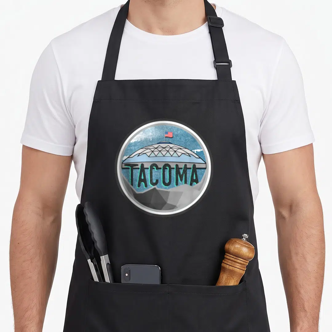 Dome tacoma apron - Circular dome illustration with TACOMA text in teal and grey, flag on top, mountains below — shown on a black cotton canvas apron against a white background