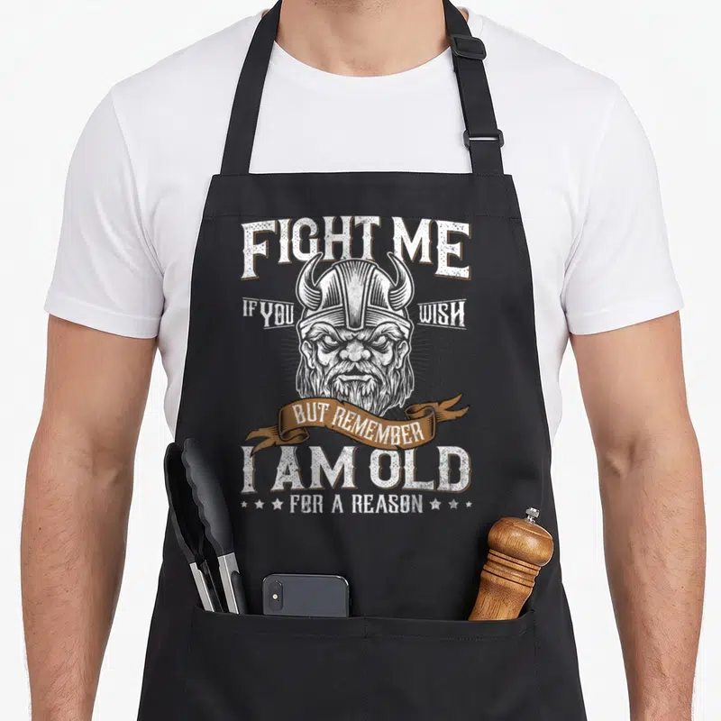 Culture viking apron - Distressed viking head with FIGHT ME quote and brown banner reading I AM OLD FOR A REASON — shown on a black cotton canvas apron against a white background