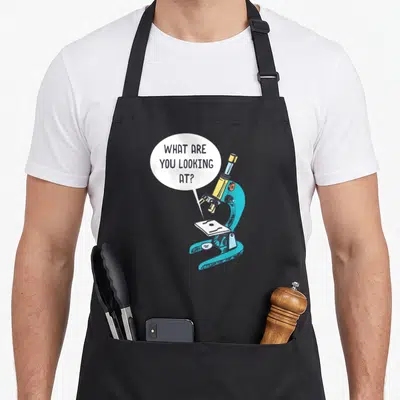 Funny microbiologist apron - Cartoon microscope with speech bubble saying WHAT ARE YOU LOOKING AT in teal and yellow — shown on a black cotton canvas apron against a white background