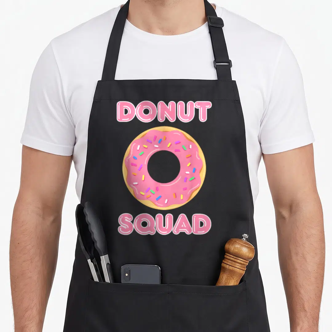 Doughnut of squad apron - Pink frosted donut with sprinkles centered between DONUT and SQUAD text — shown on a black cotton canvas apron against a white background