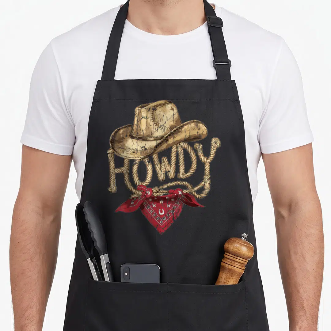 Birthday cowboy apron - tan cowboy hat above rope HOWDY text tied with red bandana — shown on a black cotton canvas apron against a white background