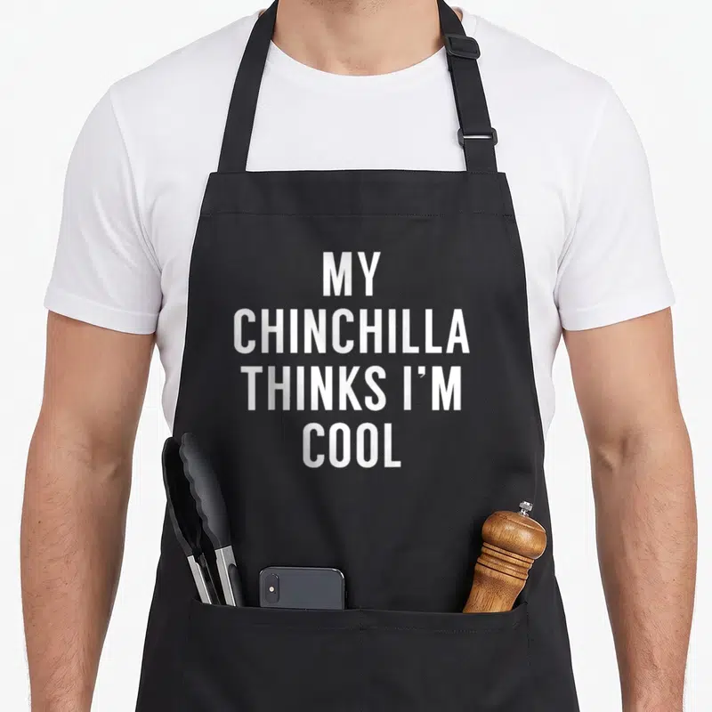 Owner crazy chinchilla man apron - White stacked text reading MY CHINCHILLA THINKS I'M COOL on a black background — shown on a black cotton canvas apron against a white background