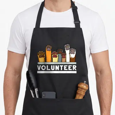Shelter grill rescue apron - Raised animal paws above white VOLUNTEER text on grey background — shown on a black cotton canvas apron against a white background