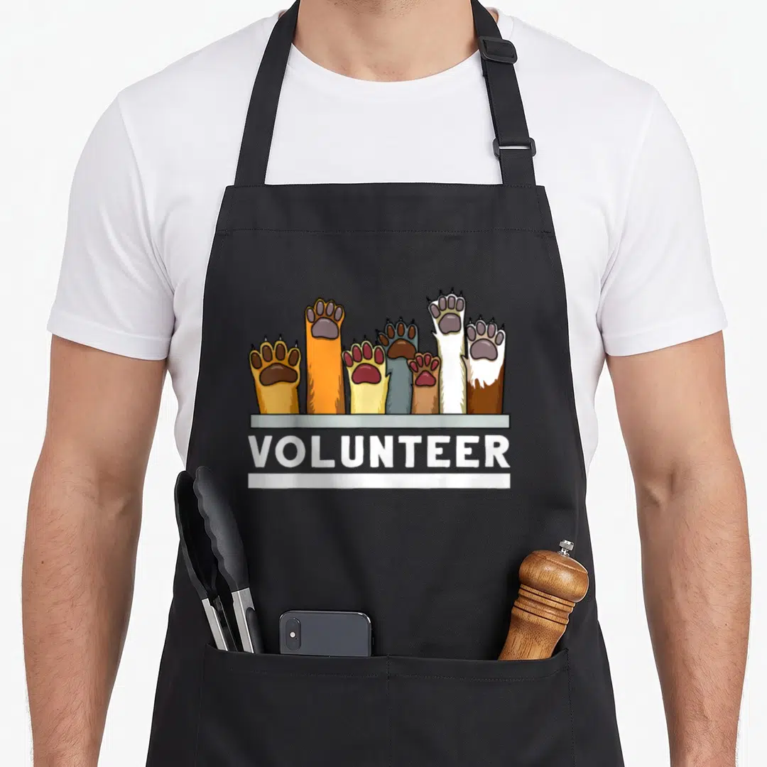 Shelter grill rescue apron - Raised animal paws above white VOLUNTEER text on grey background — shown on a black cotton canvas apron against a white background
