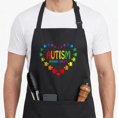 Autism april apron - Colorful puzzle heart surrounding AUTISM AWARENESS text with small red heart at bottom tip — shown on a black cotton canvas apron against a white background