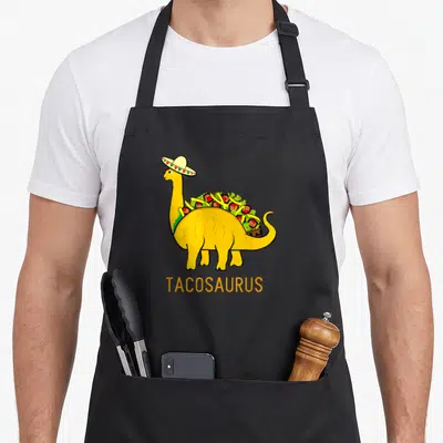 Tacosaurus dinosaur apron - Yellow dinosaur with sombrero and taco fillings, TACOSAURUS text below — shown on a black cotton canvas apron against a white background