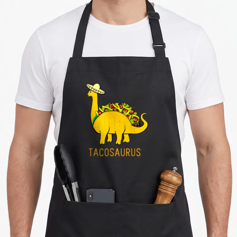 Tacosaurus dinosaur apron - Yellow dinosaur with sombrero and taco fillings, TACOSAURUS text below — shown on a black cotton canvas apron against a white background