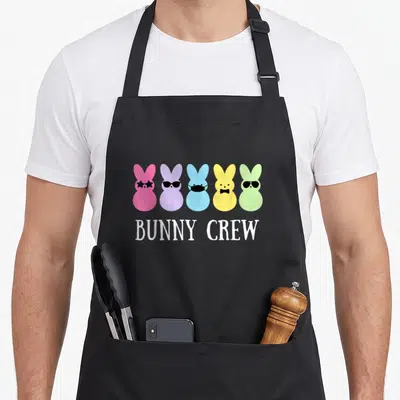 Crew easter bunny aprons - Five colorful cartoon bunny heads wearing glasses, mask, and bow tie above white text — shown on a black cotton canvas apron against a white background