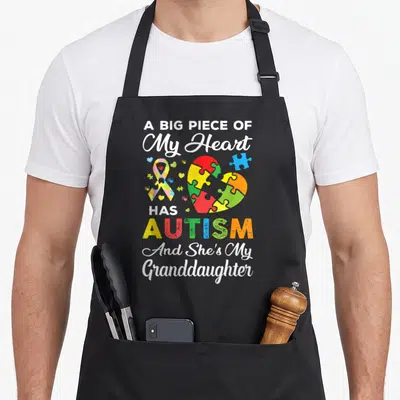 Granddaughter autism apron - Puzzle heart and ribbon with text A Big Piece Of My Heart Has Autism And She's My — shown on a black cotton canvas apron against a white background