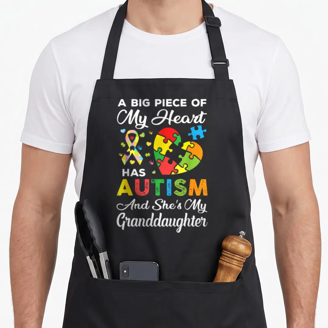 Granddaughter autism apron - Puzzle heart and ribbon with text A Big Piece Of My Heart Has Autism And She's My — shown on a black cotton canvas apron against a white background