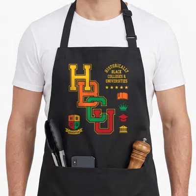 Hbcu alumni apron - Overlapping HBCU letters in yellow orange green red with shield reading EDUCATED and academic icons — shown on a black cotton canvas apron against a white background