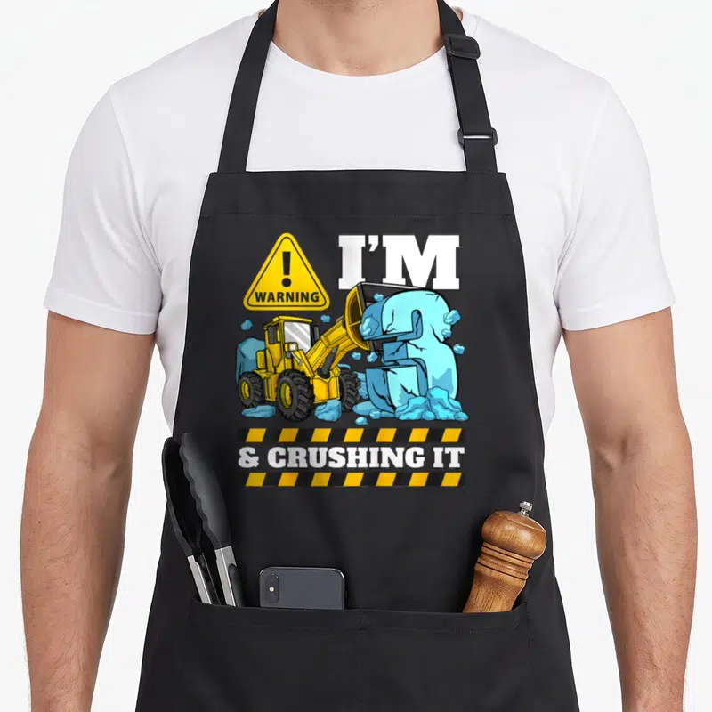 Bulldozer truck apron - Cartoon yellow loader crushing blue number 3 with warning sign and hazard stripes — shown on a black cotton canvas apron against a white background