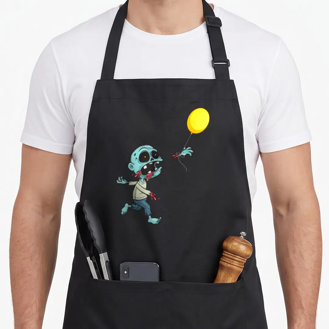 Zombie balloon apron - Cartoon zombie reaching for yellow balloon with severed hand and red blood accents — shown on a black cotton canvas apron against a white background