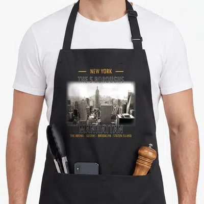Staten new york apron - Manhattan skyline photo with text listing New York's 5 boroughs in yellow and black — shown on a black cotton canvas apron against a white background