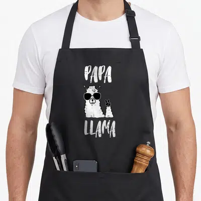 Papa llama father day apron - White text PAPA LLAMA surrounding a cartoon llama in black sunglasses on black fabric — shown on a black cotton canvas apron against a white background