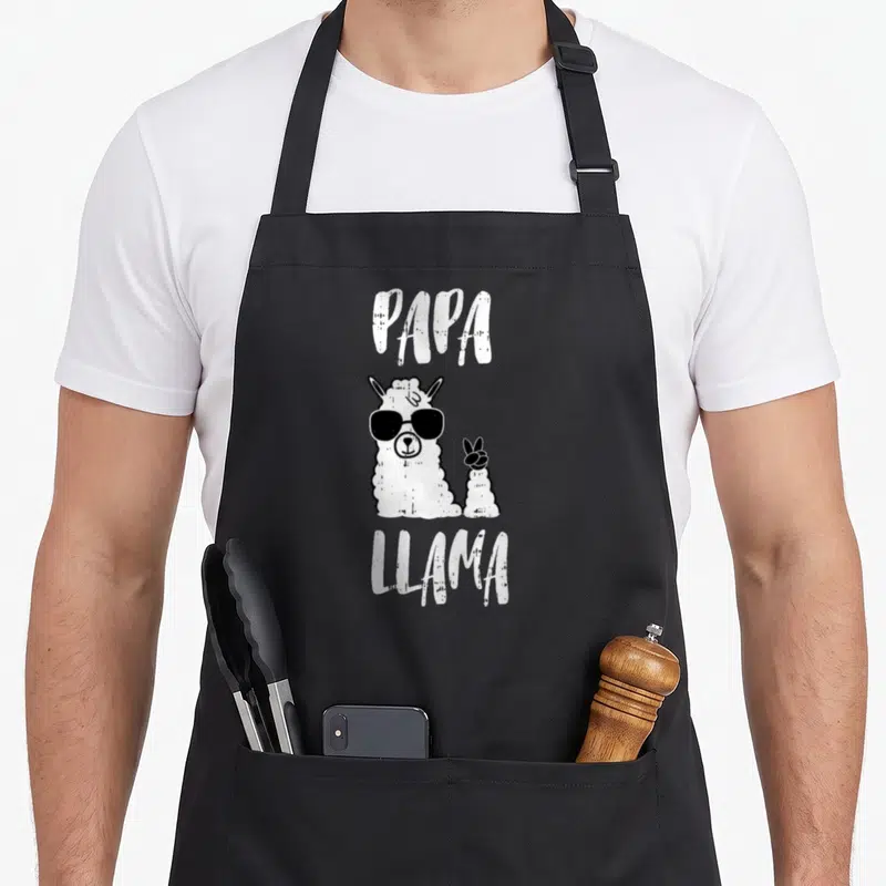 Papa llama father day apron - White text PAPA LLAMA surrounding a cartoon llama in black sunglasses on black fabric — shown on a black cotton canvas apron against a white background