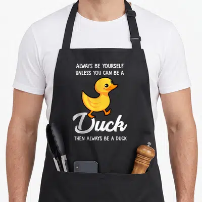 Yourself duck apron - Cartoon yellow duck walking between text Always Be Yourself Unless You Can Be A Duck Then Always — shown on a black cotton canvas apron against a white background