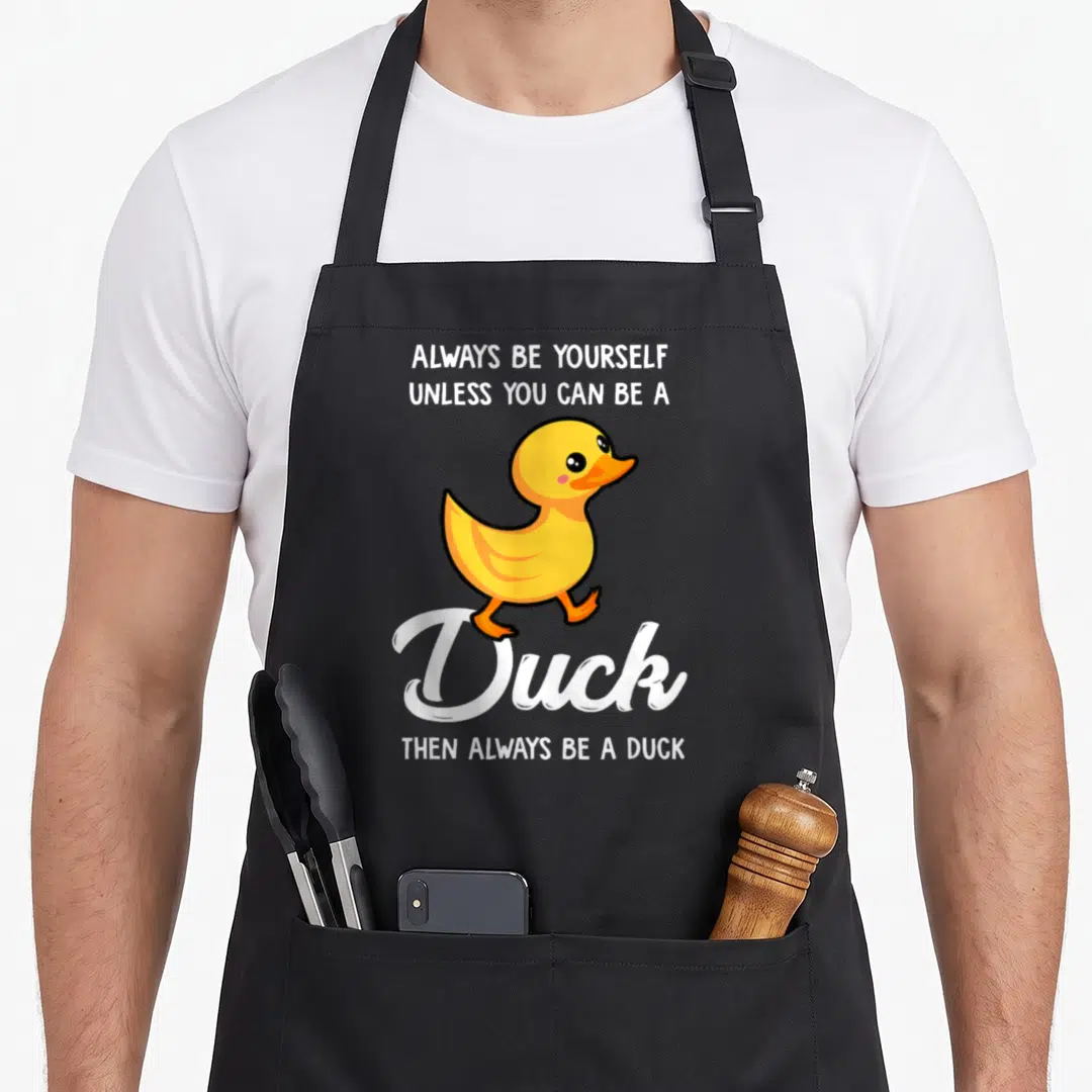 Yourself duck apron - Cartoon yellow duck walking between text Always Be Yourself Unless You Can Be A Duck Then Always — shown on a black cotton canvas apron against a white background