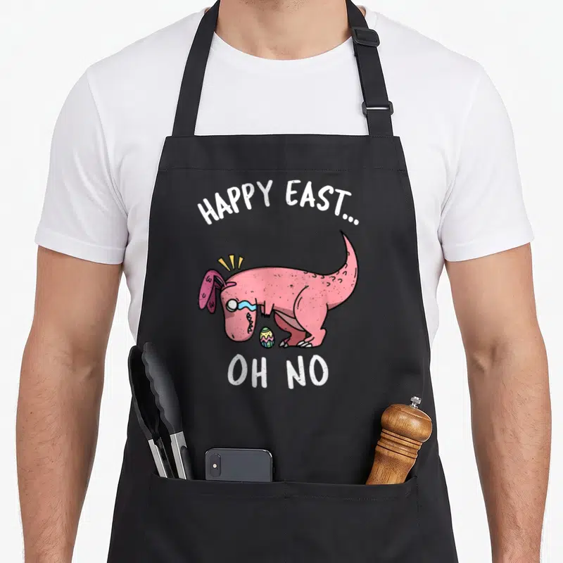 East easter apron - Pink cartoon dinosaur with bunny ears crying over egg under HAPPY EAST... OH NO text — shown on a black cotton canvas apron against a white background