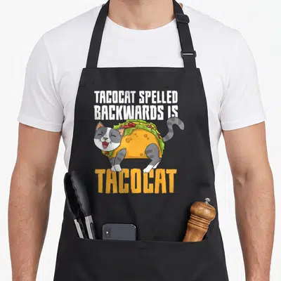 Tacocat mexican apron - Cartoon gray and white cat wearing a taco costume with palindrome text above and below — shown on a black cotton canvas apron against a white background